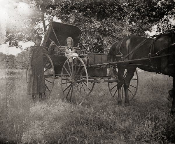 Everetta Bass sitting in horse-drawn buggy holding a doll. A girl identified as Loura is standing beside the buggy.