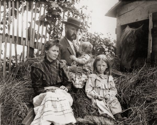 Dr. Edward A. Bass, photographer, seated in pile of hay holding his son, Cary. Seated in front of him are his wife, Ada, and daughter, Everetta. Horse identified as 'Dick' is visible in right of frame.