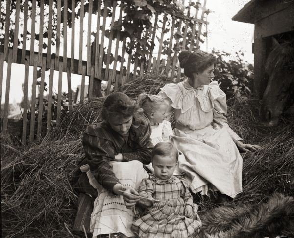 Ada, the photographer's wife, and son, Cary, seated in front of an unidentified woman and a young girl. The woman and girl are both looking at 'Dick,' the horse with his nose in the hay.