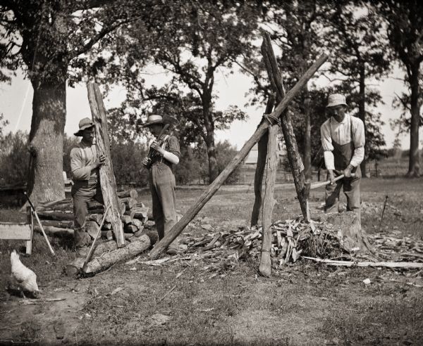 Three men cutting points on logs for fence posts.