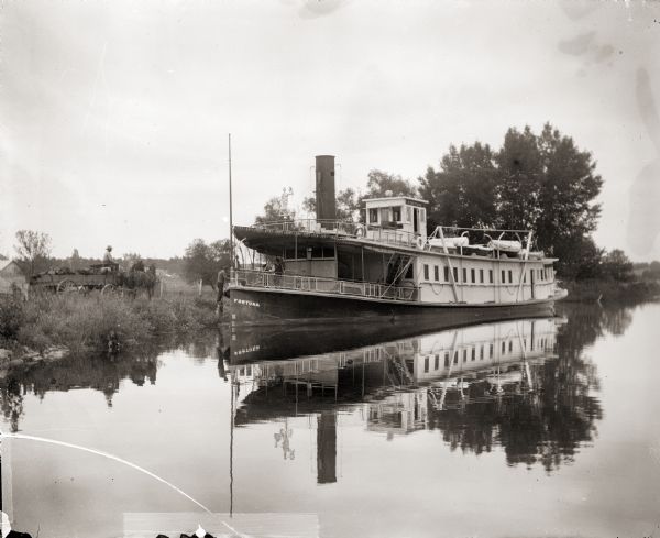 Steamboat "Fortuna" docked on the shore of an unidentified lake. A man driving a horse cart is on the shore.