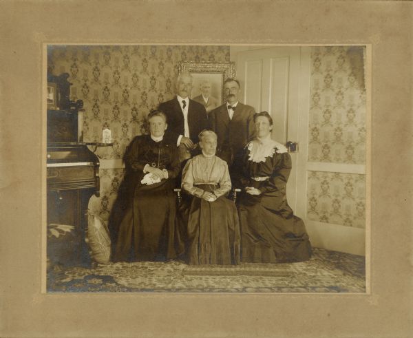 Lorinda Hill Bass sits in the center, with daughters Martha Bass Haskin, left, and Rhoda Bass Shephard beside her. Her sons, Edward, left, and Frank, right, stand behind. A portrait of her late husband, Isaac W. Bass is hanging on the wall between Edward and Frank. The photograph was taken in the parlor of the Bass farmhouse. There is a parlor organ to the left of the group.