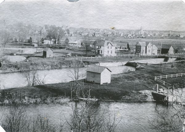 Elevated view from the south. The Fox River is in the foreground with the dam just visible in the lower right. The lock is seen in the center; the Montello River is on the left. The Methodist Church is in the left background; St. John Catholic Church is to the right.