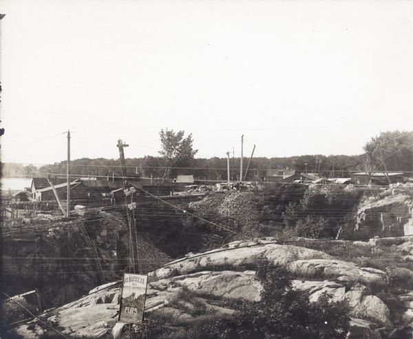 A view of the Montello granite quarry with Montello Lake in the background. The sign in the foreground reads, "J.T. Barrett Pays Highest Prices for Furs, Montello."