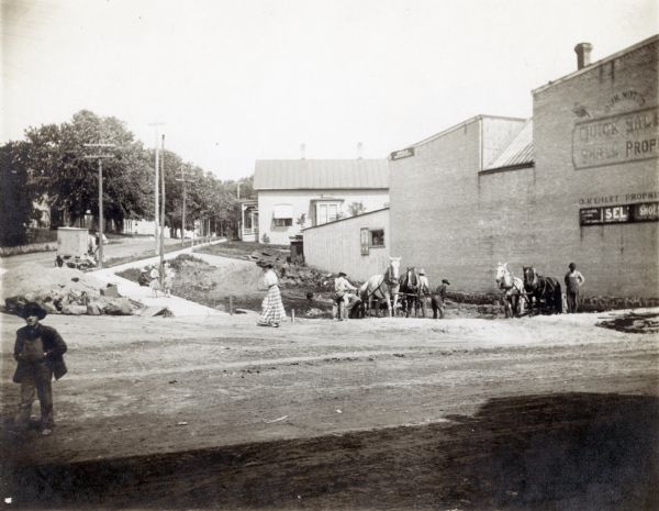 Workmen with teams of horses excavate for the new Mercantile Store, later the M.M. Smart Store. A woman is walking on the street nearby, and a boy or man wearing overalls is on the left.