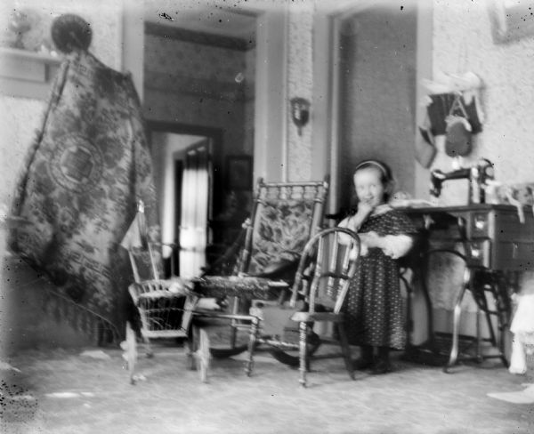 Everetta Bass stands in a room near chairs in front of a treadle sewing machine. A doll buggy is also visible. A tapestry or blanket hangs on the wall on the left.