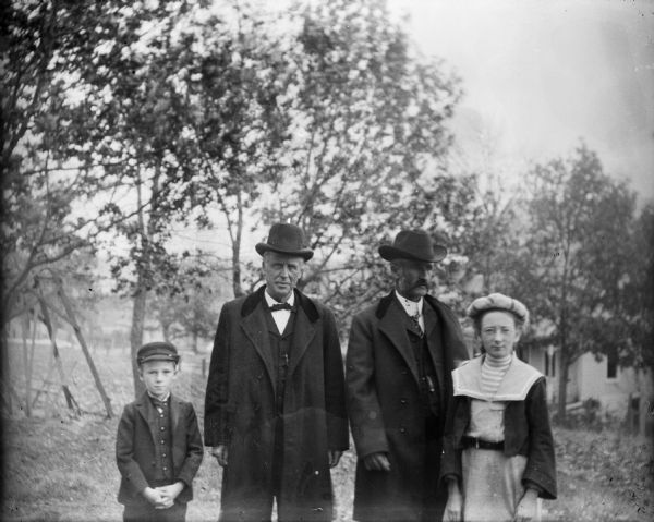 Dr. Bass, second from right, with his uncle, Rev. Edward Cary Bass, and Dr. Bass's children Edward Cary and Everetta. They are standing outdoors with a lawn swing in the background.  The house visible on the right was built by C.F. Roskie in 1902.
