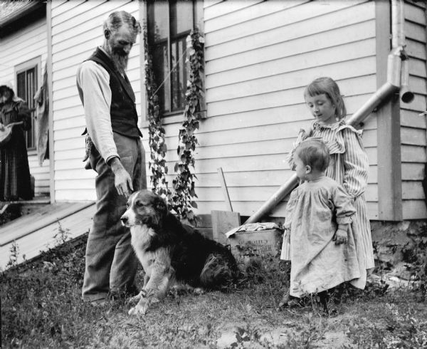 The photographer's father-in-law, Leroy J. Burlingame, with Everetta and Cary Bass and the dog, Cuff in the yard of a house.  An unidentified woman stands in the background on a porch of the house.