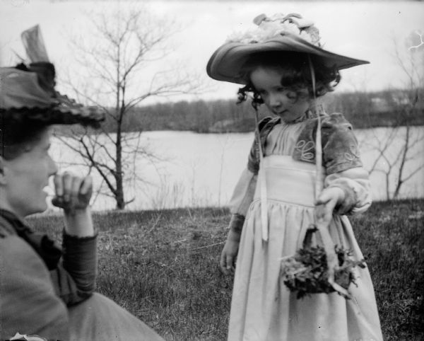 Everetta Bass, holding a basket of flowers, and her mother Ada on a hill by the mill pond (Montello Lake). The far shoreline of the lake can be seen in the background. A painting of Everetta in this pose is featured in several later Bass photographs.
