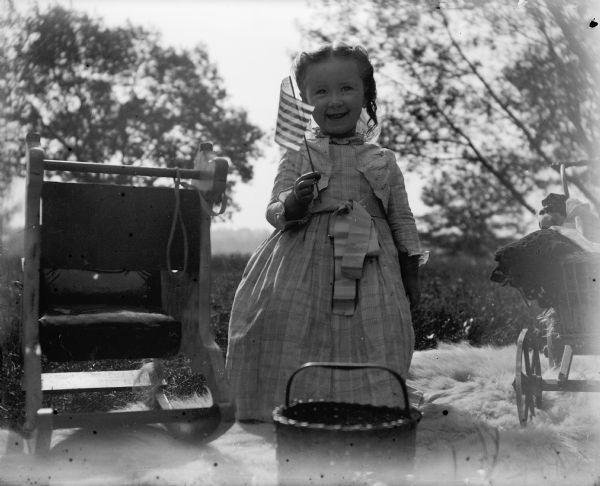 Everetta Bass stands on a fur rug outdoors holding a small American flag. A rocking horse, basket, and doll buggy are nearby.
