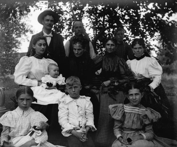 A group of unidentified men, women and children photographed on a bentwood bench outdoors. One girl holds a doll, and a woman holds an infant.