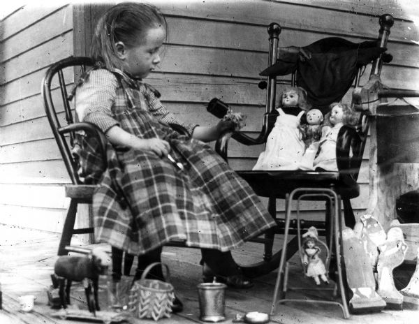 Everetta Bass, sitting in a child's chair, holds a goblet and spoon in the company of her dolls and other toys on the porch of the Bass house.