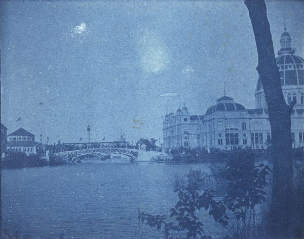A cyanotype view across lagoon from shoreline of the U.S. Government Building (right), and the Fisheries building (left). There is a bridge over the lagoon between the two buildings.