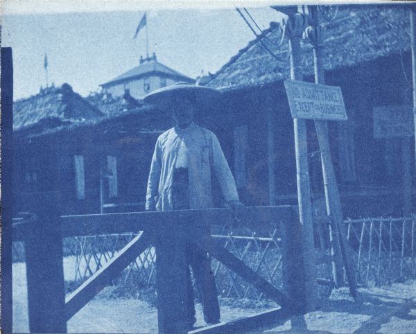A cyanotype photograph of a man in oriental dress on the Midway of the World's Columbian Exposition. He stands behind a gate; a sign warns, "No Admittance Except on Business".  There is a low rustic fence and several thatched buildings in the background.