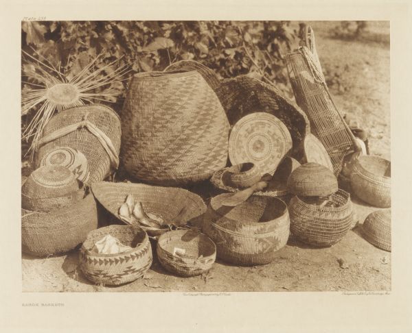 The original caption reads: "The basketry of the Karok does not differ from that of the Hupa and the Yurok. The process is always twining, and the usual materials are hazel rod for the warp, roots of the digger or the yellow pine for the weft, and Xerophyllum grass for white overlay, bark of the maidenhair fern for black, and fibres from the stem of Woodwardia fern, dyed in alder-bark juice in the mouth of the workwoman, for red. Represented in the plate are the receptacle for the storage of seeds and nuts, the burden-basket, the winnowing tray, various sizes of mush-baskets and food containers, and the cradle-basket." A view of several different type of baskets.