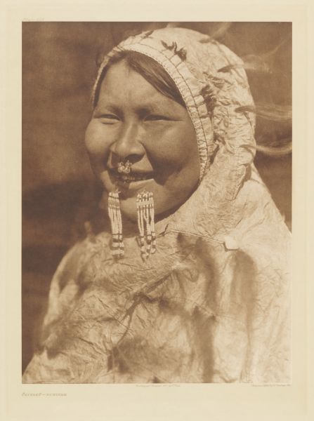 The original caption reads: "This contented young woman wears a nose-ring and a labret. Her waterproof hooded parka is made of intestinal parchment." A head and shoulders portrait of a woman with nose piercing.