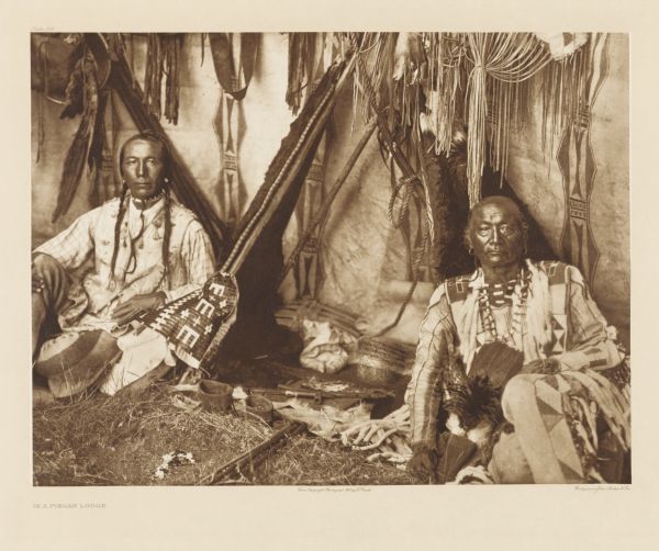 The original caption reads: "Little Plume with his son Yellow Kidney occupies the position of honor, the space at the rear opposite the entrance. The picture is full of suggestion of the various Indian activities. In a prominent place lie the ever-present pipe and its accessories on the tobacco cutting-board. From the lodge-poles hang the buffalo-skin shield, the long medicine-bundle, an eagle-wing fan, and deerskin articles for accoutering the horse. The upper end of the rope is attached to the intersection of the lodge-poles, and in stormy weather the lower end is made fast to a stake near the centre of the floor space." Indoor portrait of two seated men. The original glass negative also captured a box on the ground between the two men, which contained a clock or a metal. The copper-plate was retouched to remove the item.