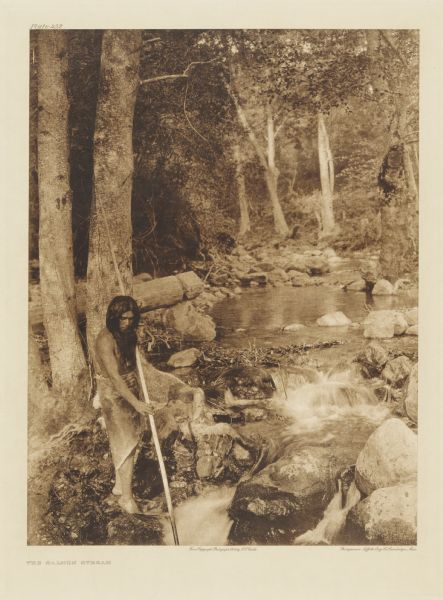 The original caption reads: "A Hupa youth is waiting with poised spear for the shadowy outline of a salmon lurking in a quiet pool and gathering its strength for a dash through a tiny cascade." A view of a man fishing with a spear in a river.