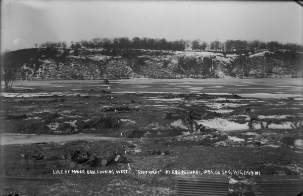 Frozen Wisconsin River from the east bank looking west, marked to show the future location of the power dam.  Logs and stumps indicate prior clearing of the east shore.  Pilings are stacked in the foreground.