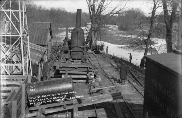 Workers erecting a steam driven pile driver while others survey for the railroad extension from the village of Prairie du Sac to the dam site on the Wisconsin River.
