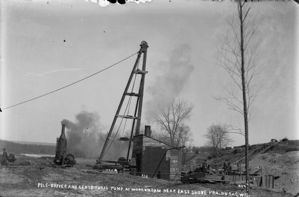 Steam-powered pile driver and centrifugal pump (in shed) at work at the Wisconsin River dam site. Men and children are watching the work.