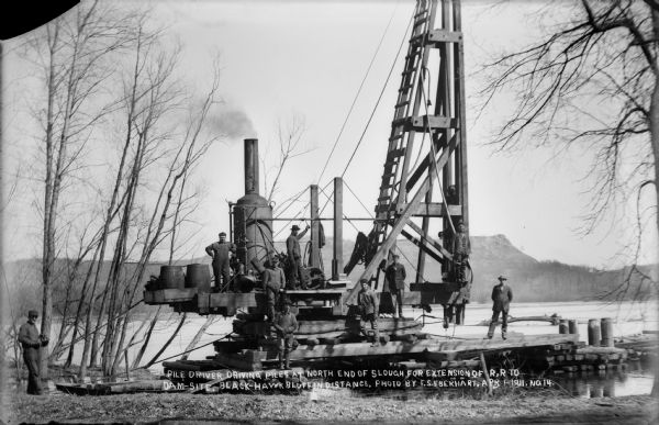 Workmen pose on a steam-powered pile driver at work at the north end of the slough on the Wisconsin River. The railroad was extended from the village to the dam site at the time of construction. Blackhawk bluff is in the distance.