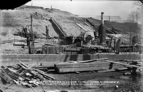 Newly poured concrete abutment on the east shore of the Wisconsin River. Iron reinforcing rods protrude from the concrete. Workmen and the steam-driven cement mixer are also visible.