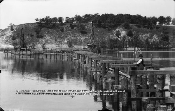 Looking west over the Wisconsin River at the unfinished narrow gauge railroad trestle built for construction of the dam. Two pile drivers are seen in the area of the coffer dam. A man is taking in the view from the trestle.