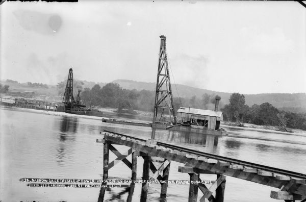 Looking east over the Wisconsin River showing the unfinished narrow gauge railroad trestle. Two steam-driven pile drivers on barges with construction workers are at work on the coffer dam which will allow construction of the power house. Bluffs are visible on the east shore.