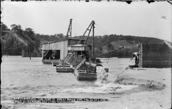 A steam-powered centrifugal sand pump at work on the Wisconsin River during the construction of the power dam. Pilings of the coffer dam are visible in the background. Nearby, a man is working on a wooden form.