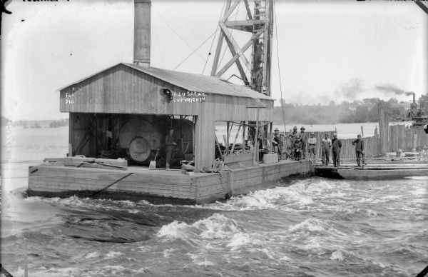 Workmen stand on a barge carrying a steam-powered pile driver. The pilings will form the coffer dam necessary for construction of the power plant on the Wisconsin River.
