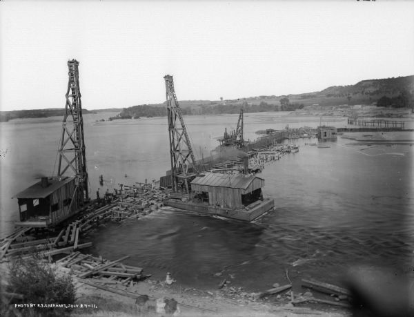 Elevated view of the power dam construction site on the Wisconsin River from the west shore. The outline of the coffer dam is easily seen.  Three steam-powered pile drivers are at work. The small shed in the distance contains the centrifugal sand pump. Spectators on shore watch the men and machinery at work.