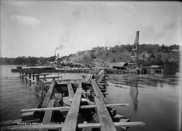 Dam site on the Wisconsin river, looking southwest along the unfinished narrow gauge railroad trestle. The coffer dam and two pile drivers are also visible. Men are at work on the site.
