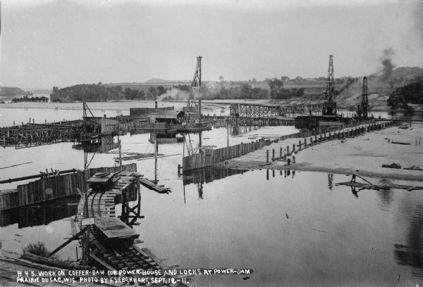 An overview of the dam site on the Wisconsin River from the west shore.  Three pile drivers are at work on the coffer dam.  The railroad trestle is in the foreground; the middle rail carried the electric current to power the train.  The centrifugal sand pump is in the shed-like structure on the far left.