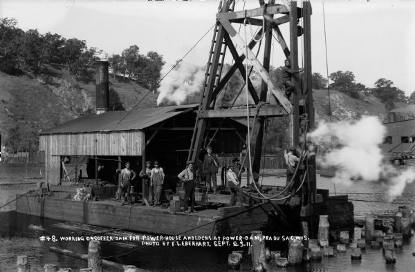 Workers standing on the barge-mounted, steam-driven pile driver at work on the coffer dam.