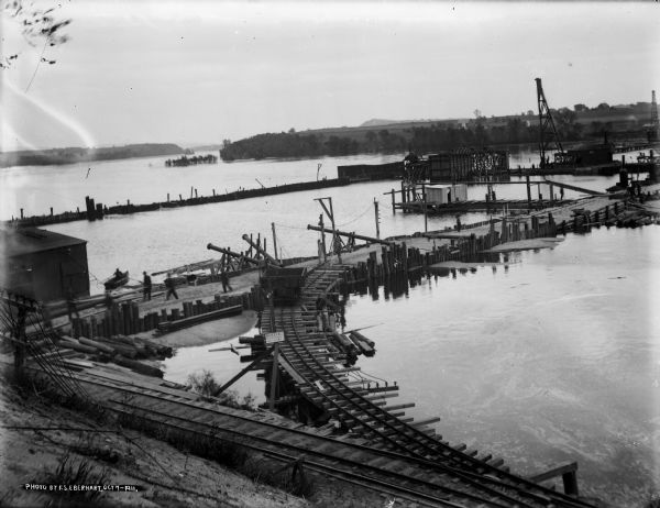 Elevated view of the rising waters of the Wisconsin River at the construction site. This view shows the coffer dam, pile drivers, and narrow gauge railroad. Note the partially submerged trees upriver.