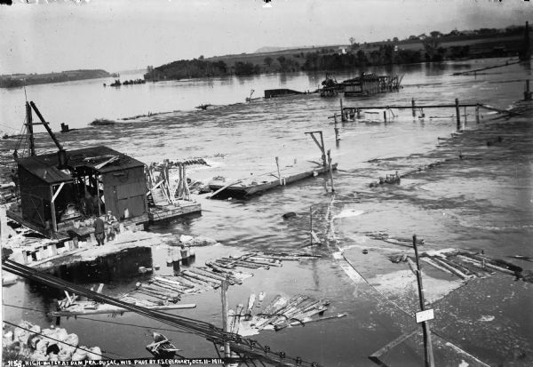 Elevated view of high water on the Wisconsin River which has inundated the coffer dam at the construction site. The narrow gauge railroad (curved line in foreground, right) is nearly submerged. Two workmen are standing behind one of the pile drivers.