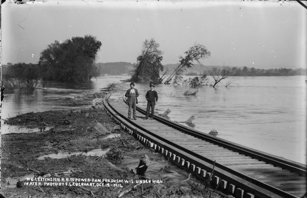 Two men are standing on the tracks of the newly constructed railroad extension to the construction site of the Prairie du Sac dam.  Behind them, the tracks are submerged in floodwaters of the Wisconsin River.