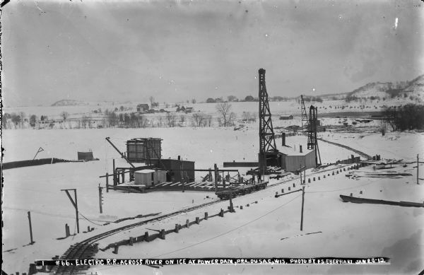 Elevated view of two men standing on a rail car on the narrow gauge electric railroad crossing the river at the dam construction site. The portion of track in the background is at the level of the ice.