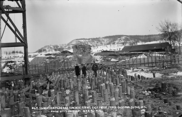 Two women and a man posing among the timber pilings near the east shore of the river. Wooden concrete forms and two buildings are in the background.