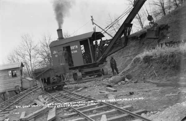 Workmen operating a steam shovel loading gravel into electric rail cars during construction of the power dam.