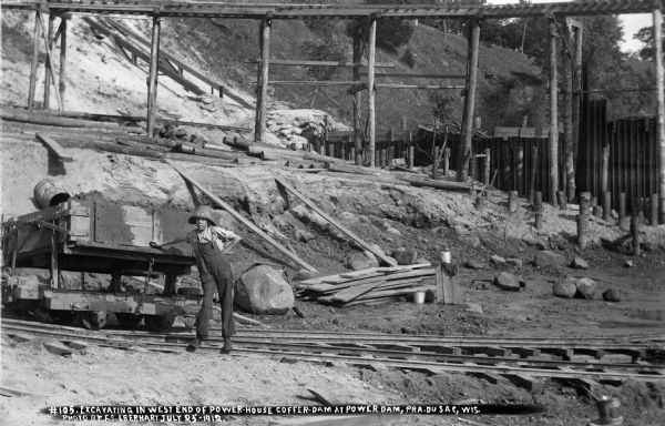 A boy posing in the excavation site for the power house within the coffer dam. He is standing on the narrow gauge tracks; the high trestle is in the background.