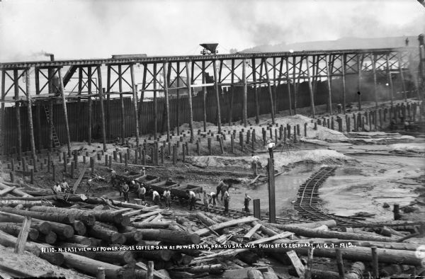 Men excavating within the coffer dam. One man is standing on a high piling. Several steam engines at work fill the air with smoke. The high trestle and foundation pilings are easily seen.