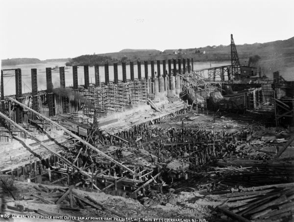 Elevated view of the progress of construction within the coffer dam showing the gate guides and draft tubes in place. Concrete forms and chutes, as well as timber pilings, are also visible.