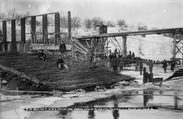 A winter view with workers posing with brush mats at the power dam construction site. The gate guides and high trestle form the background.