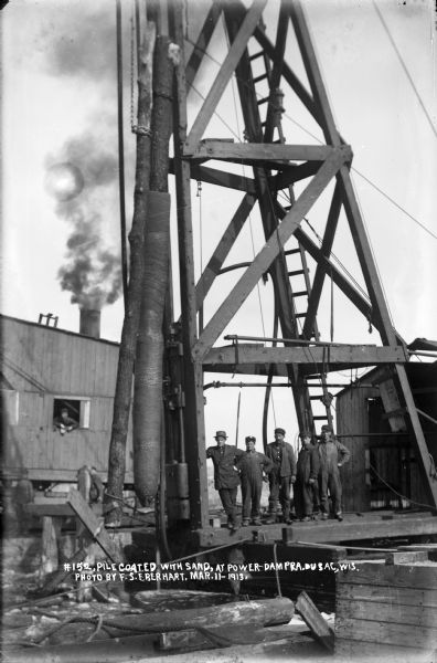 Workers posing by a piling coated with sand suspended from a crane.