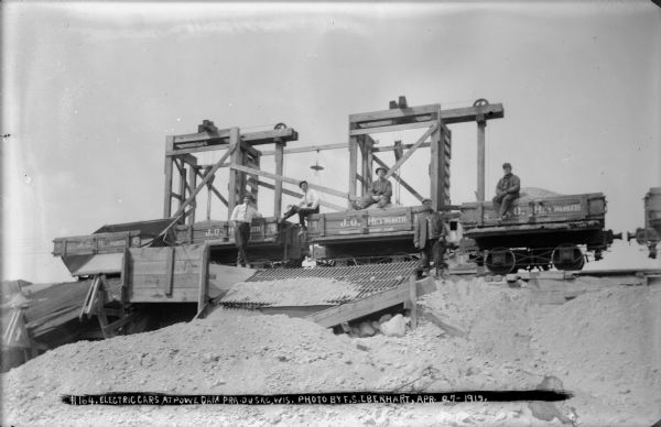 Five men, two well-dressed, posing on electric rail cars during construction of the dam.