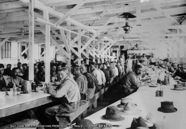 Power dam construction workers pausing during a meal in the camp dining room. Oil lamps are hanging from the ceiling; overhead electric lights are also installed.