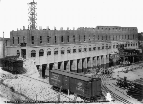 Elevated view of the power house from the southwest showing progress of the brickwork. Two railroad cars are standing on the railroad extension. Workers are pushing cars along the narrow gauge track.
