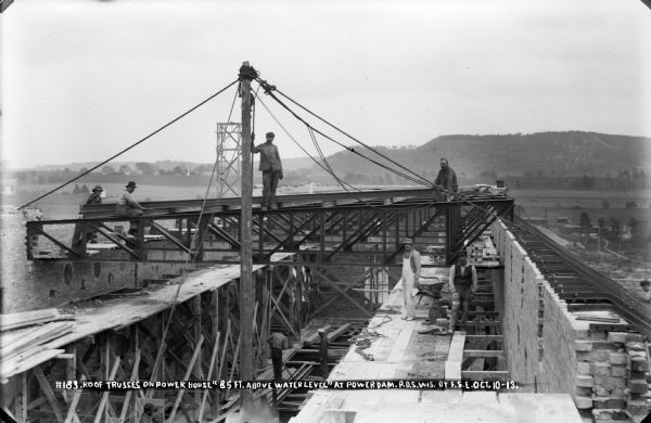 Workers posing with roof trusses on the power house, 85 feet above the water level at the Prairie du Sac Dam. Note the brickwork in progress.